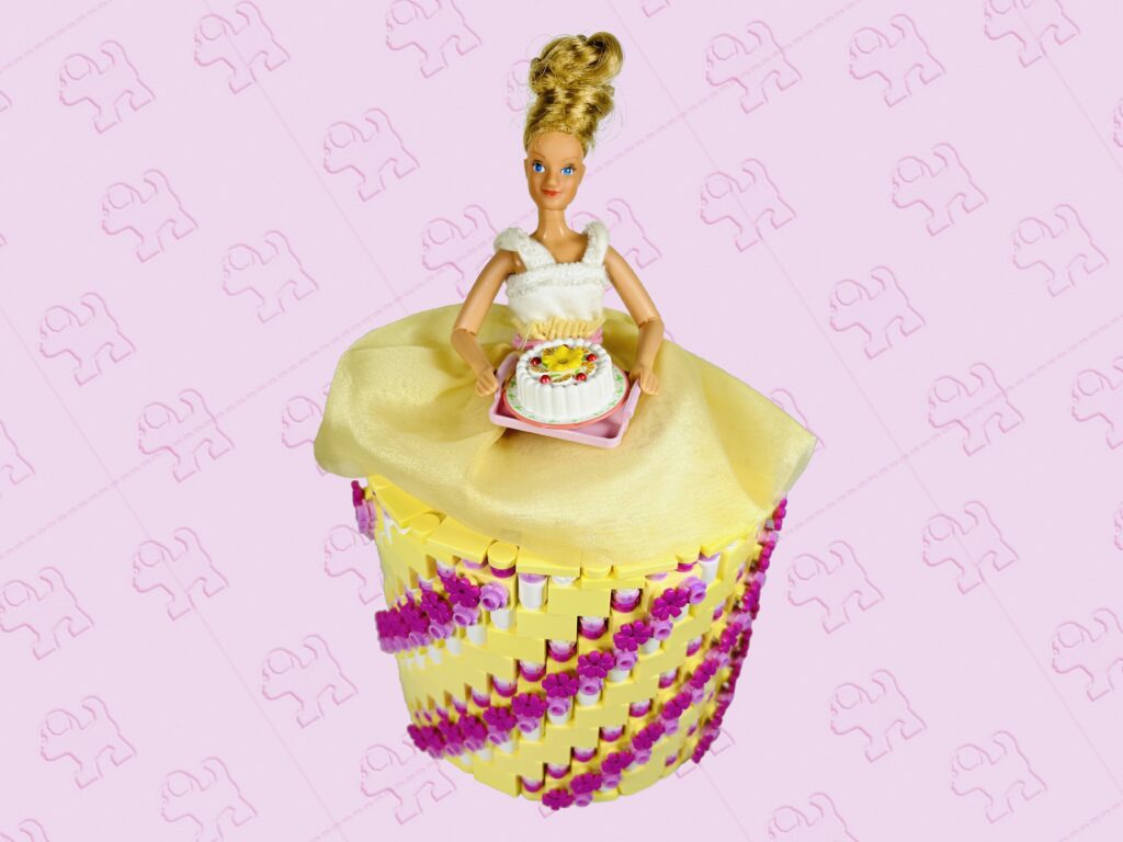 Blonde doll carrying a cake on a tray. She’s wearing a bright light yellow, bright pink and dark pink coloured brick-built toilet roll dress.