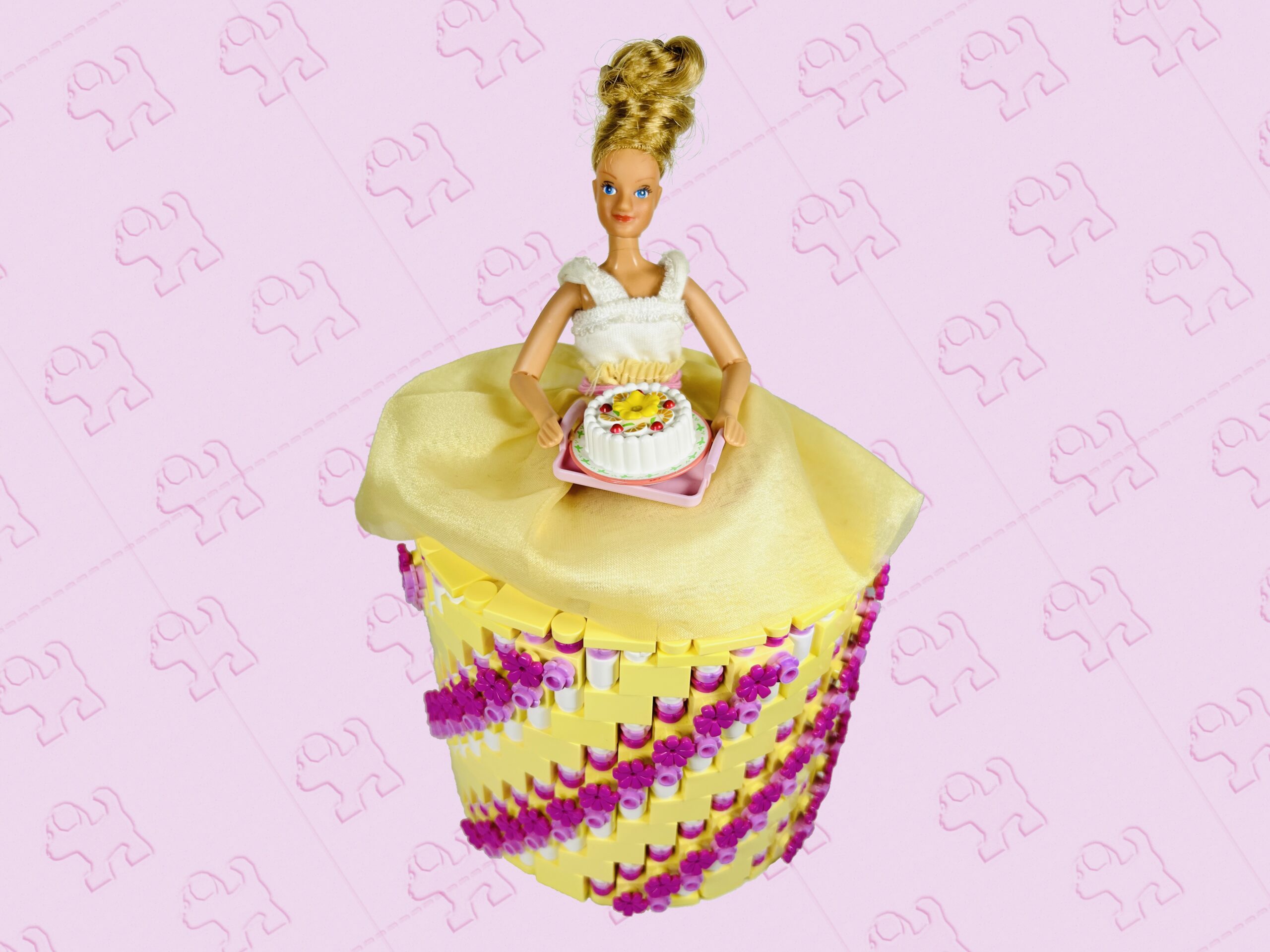 Blonde doll carrying a cake on a tray. She’s wearing a bright light yellow, bright pink and dark pink coloured brick-built toilet roll dress.
