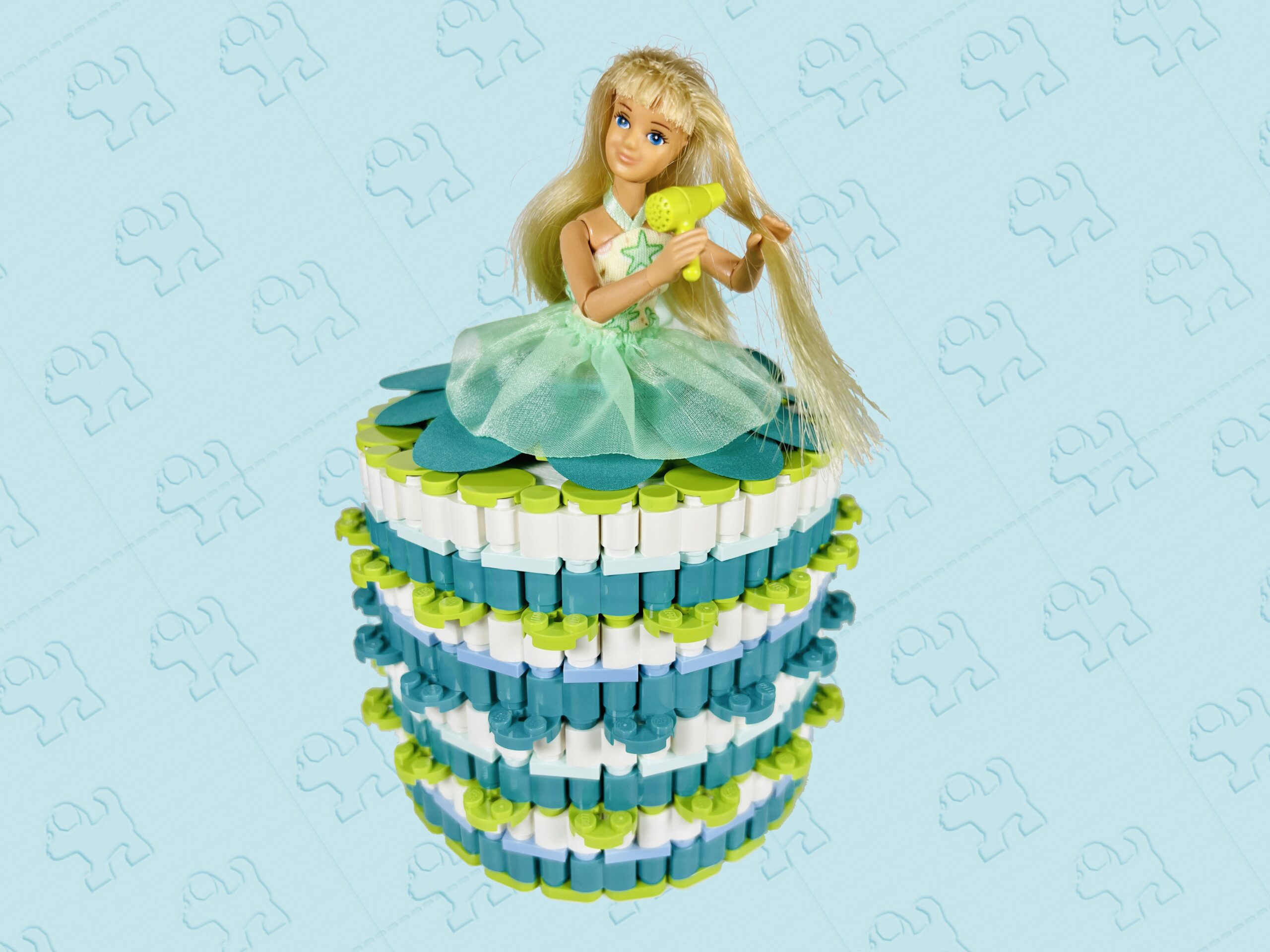 Blonde doll drying her hair. She’s wearing a white, lime and dark turquoise coloured brick-built toilet roll dress.