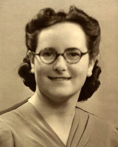 Sepia photograph of a young Peggy smiling with round glasses and dark hair in a smart symmetrical pin curl style.