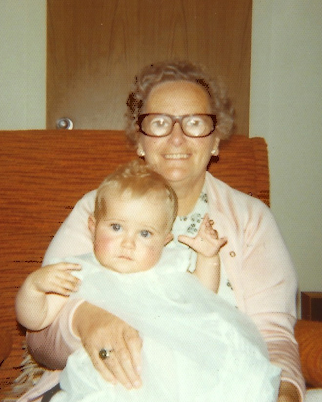 Polaroid of smiling woman, sitting down, holding a young baby in a white gown.