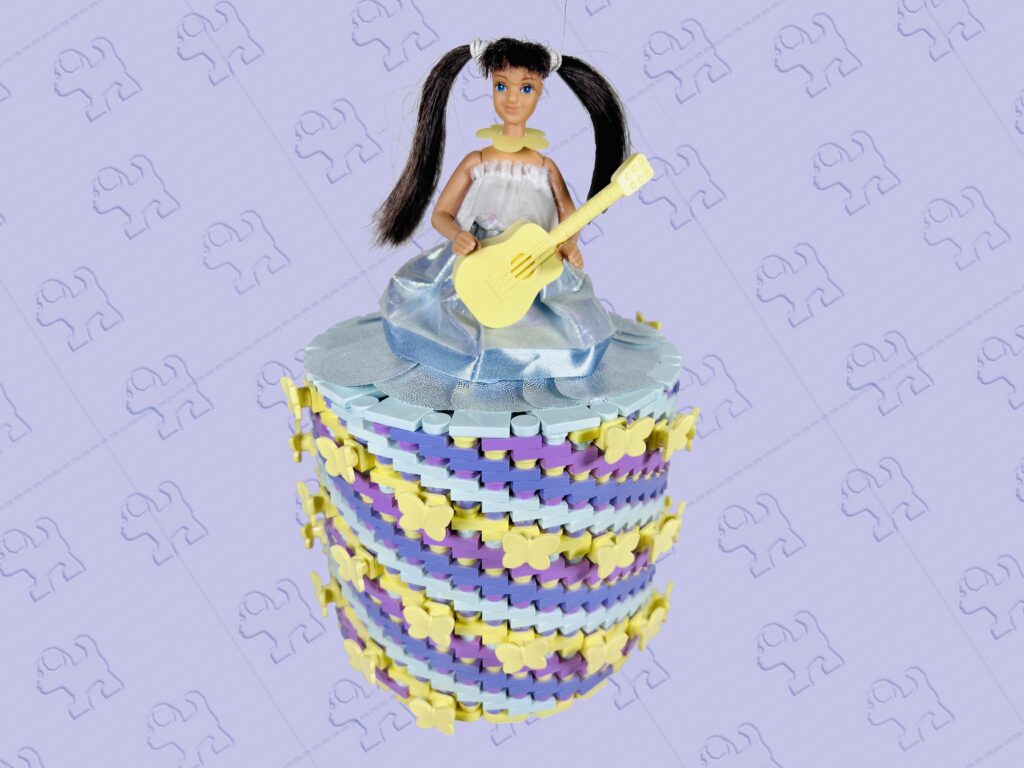 Brunette doll playing an acoustic guitar. She’s wearing a light aqua and bright light yellow coloured brick-built toilet roll dress.