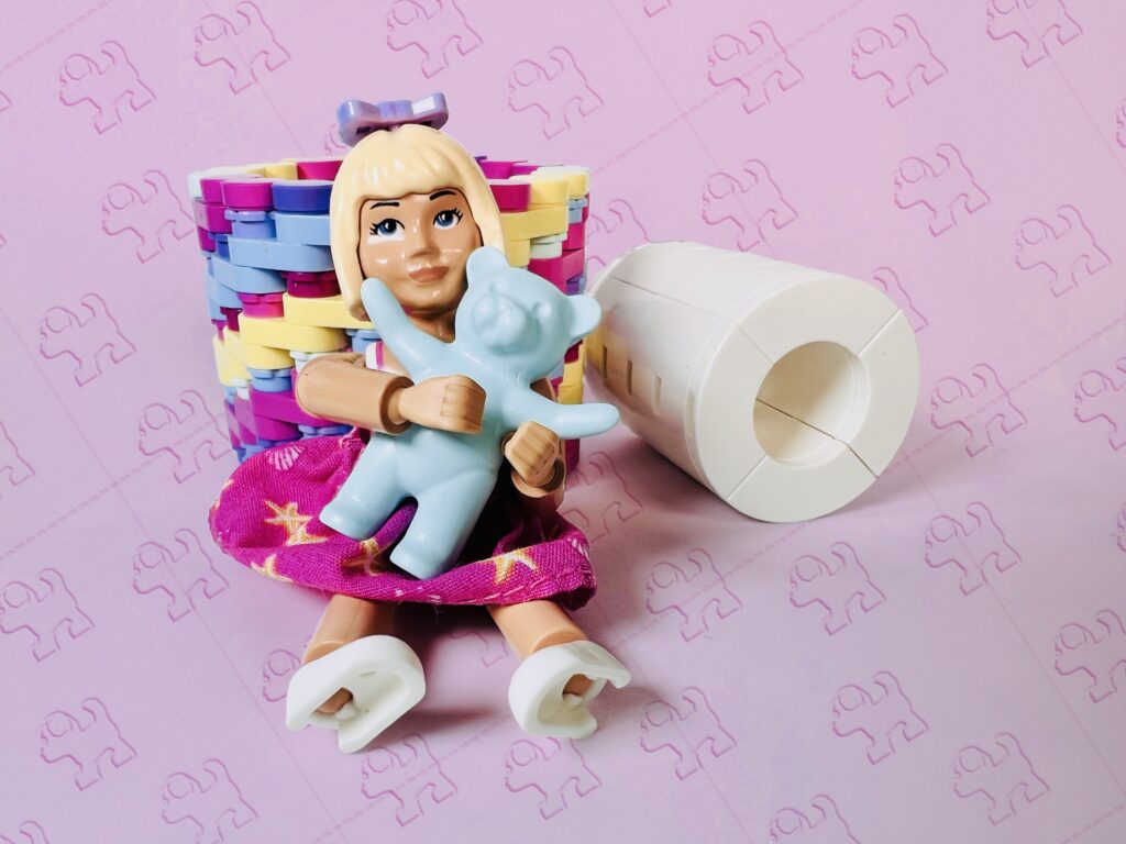 Small blonde doll holding a teddy bear, next to a brick-built toilet roll.
