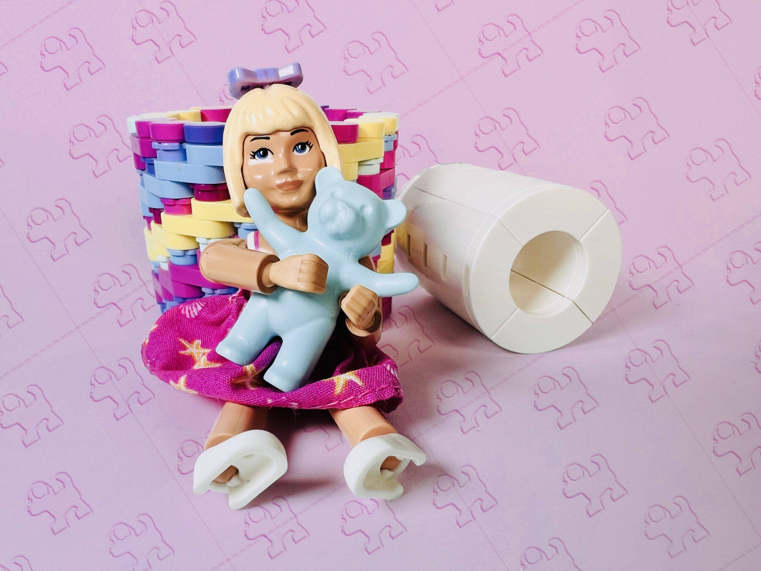 Small blonde doll holding a teddy bear, next to a brick-built toilet roll.