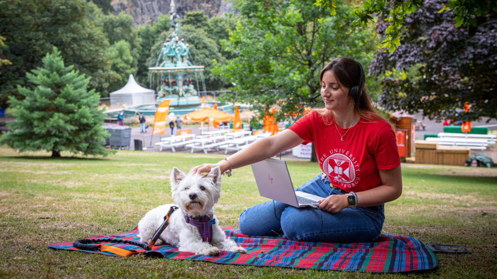 Young student using a laptop, sitting in a pretty park with her cute white dog.