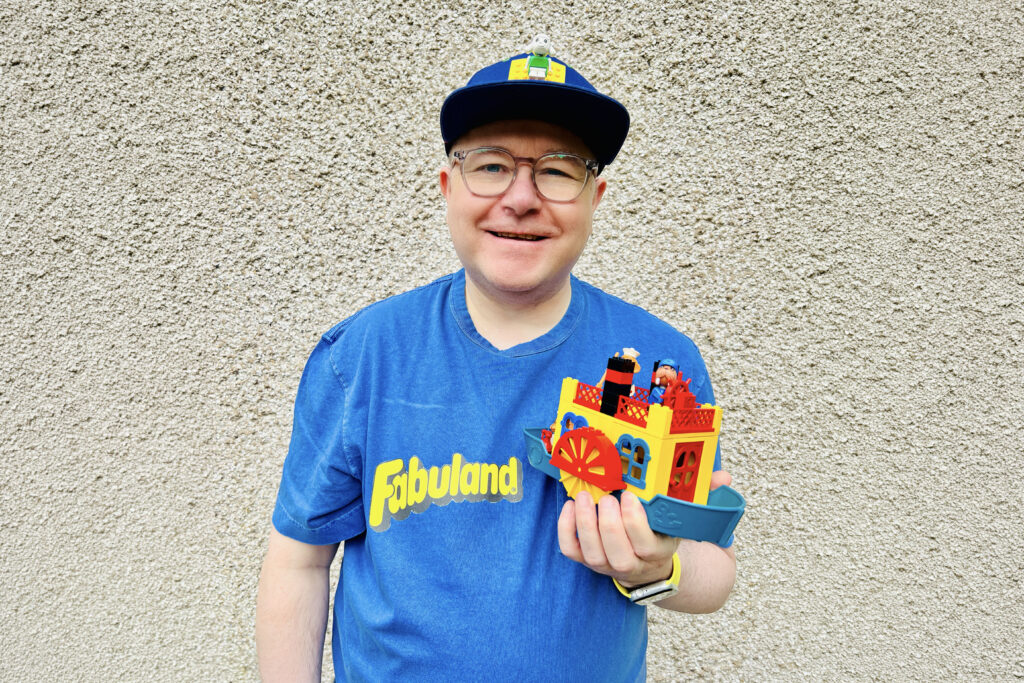 Stewart smiling, wearing a blue Fabuland T-shirt holding a small, colourful Lego Fabuland boat in front of a textured beige wall.