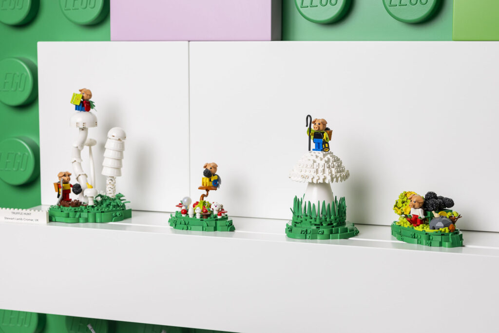 Four small Lego vignettes labelled “Truffle Hunt" featuring minifigure pigs in mushroom-like structures, arranged on a white shelf against a background of large green bricks.